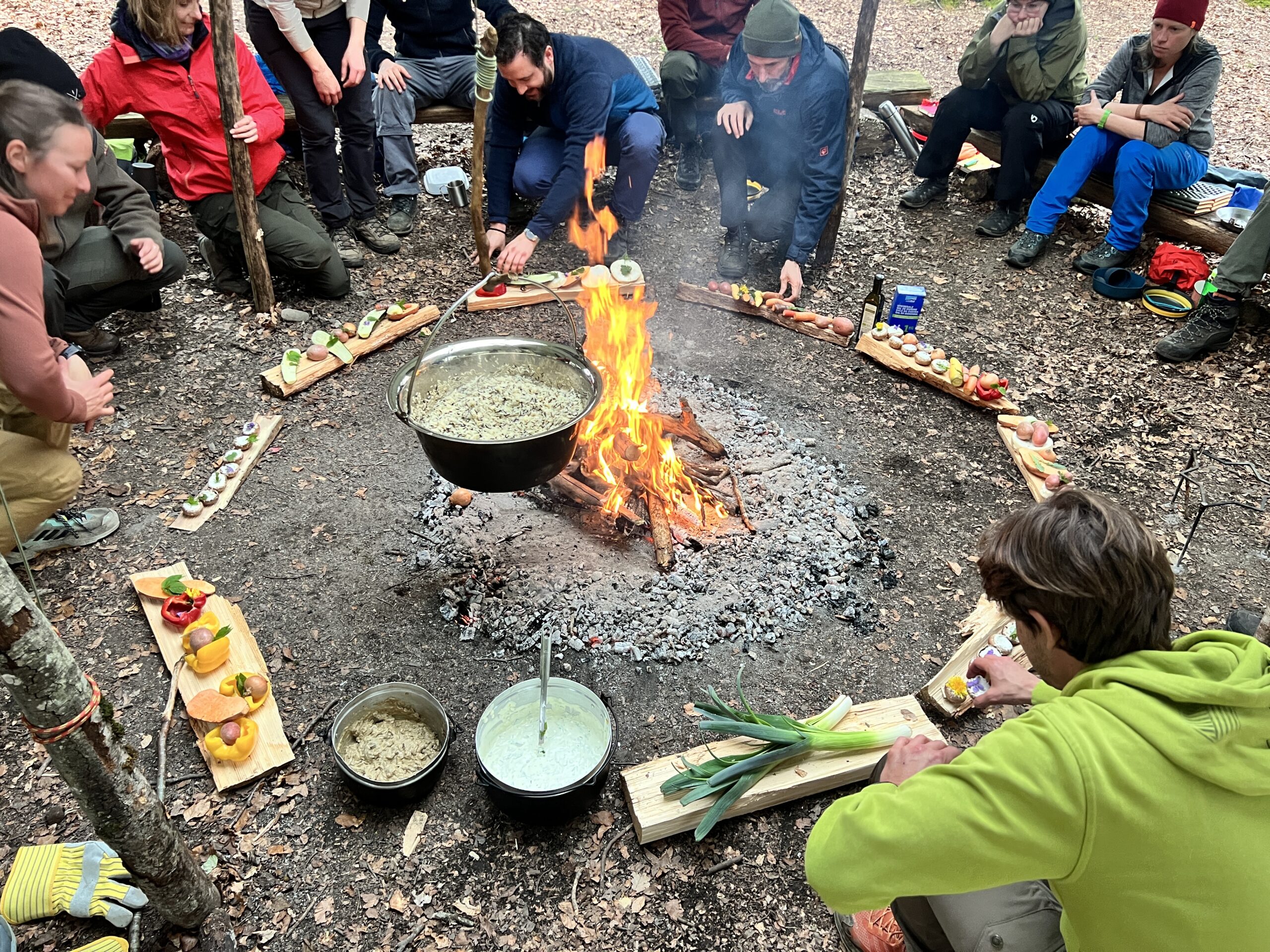 Gruppe am Lagerfeuer mit hängendem Kochtopf und Holzplatten voller Snacks